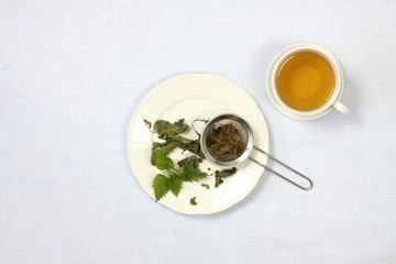 Healing tea with fresh nettles, flat view. Cup of nettle tea , dry and fresh nettles, tea strainer on the plate,  light tablecloth