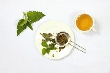 Healing tea with fresh nettles, flat view. Cup of nettle tea , dry and fresh nettles, tea strainer on the plate,  light tablecloth