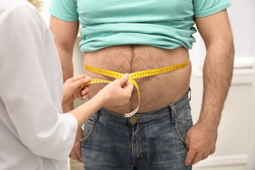 Doctor measuring overweight man's waist in hospital, closeup