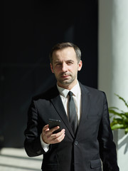 Waist-up portrait of middle aged businessman in formal black suit and necktie  standing in office hall looking at camera holding cell phone in his hand