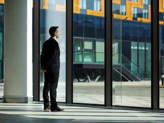 Fototapeta premium Confident businessman in black suit standing in office entrance hall with hand behind back and looking through window, thinking of new ideas, planning project or waiting for somebody to come