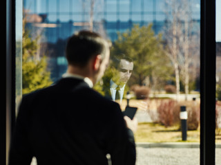 Back view of middle aged businessman standing by window in office and using cell phone, focus on his reflection in glass