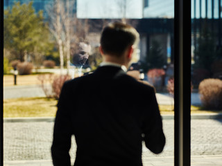 Rear view of businessman standing near window in office and text messaging on cell phone, focus on his reflection in glass