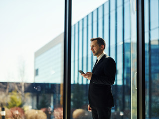 Side view of confident manager in black suit standing by floor length window, holding mobile phone and looking away thoughtfully
