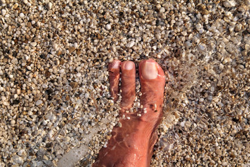 Top view of man standing bare feet on beach. Texture of bottom, leg and foot of man drowning with sweeping colorful stone below on rocky stones beach in sea with waves and foam, water ripples of sea.