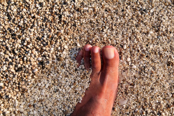 Top view of man standing bare feet on beach. Texture of bottom, leg and foot of man drowning with sweeping colorful stone below on rocky stones beach in sea with waves and foam, water ripples of sea.