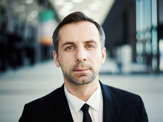 Close up portrait of confident middle aged businessman in suit, white shirt and necktie looking at camera in office lobby, stubble on his face