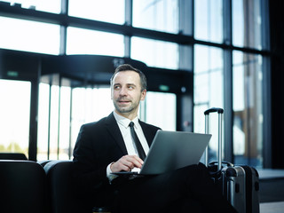 Handsome businessman looking away at somebody while sitting on leather sofa in hotel lobby or airport lounge and working on laptop, his trolley suitcase placed nearby