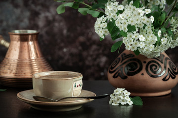 On the table is a cup of coffee and a vase with white flowers of spirea