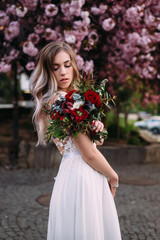 stylish and beautiful young bride dressed in a wedding dress, stands near a tree in her hands holding a bouquet