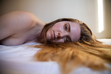 Portrait of young lonely naked pretty woman with brown hair lying on bed and looking at camera