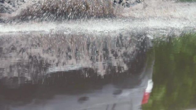 Close-up slow motion shot of car tyre driving over puddle with dirt water and splashing across wet road after rain. Rain in rush hour - Car driving on urban roads in rainy weather.