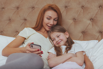 Lovely happy mother and her cute little daughter using smart phone at home together. CHeerful little girl smiling, her mother showing her something online on mobile phone, copy space