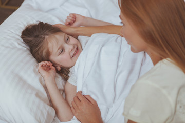 Cute cheerful little girl laughing joyfully, lying in her bed. Adorable happy girl waking up , smiling at her mother. Woman waking up her child in the morning. School, home, childhood concept