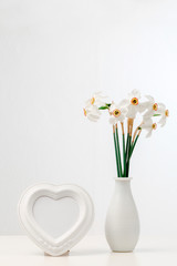 Mockup with white frame in the shape of a heart. Daffodils in a white vase on a white table. Home interior with decor elements.