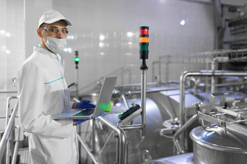 Portrait of man in a white robe and a cap standing in production department of dairy factory with laptop