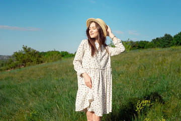 Half length portrait of a charming positive woman dressed in long white summer dresses with a straw hat of happy smiles against the background of a green meadow and blue sky. Beautiful woman enjoying