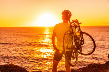 Triathlon road bike cyclist carrying bicycle watching sunset after outdoor race training by ocean coast landscape. Man athlete biking outdoors.