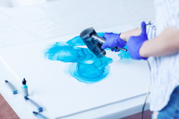 Hands of unrecognizable female artist in gloves using hair dryer while creating fluid acrylic...