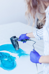 From above view of unrecognizable female artist in gloves using hair dryer to create turquoise fluid acrylic painting on canvas