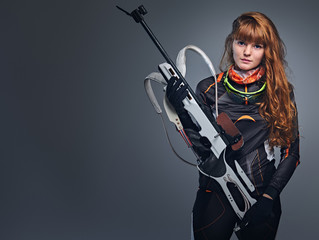 Studio portrait of a redhead female Biathlon champion holds a gun over grey background.