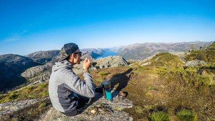 A young man preparing a dinner in the wilderness on a portable camping stove, with a view on the...