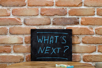 Chalk board with the phrase WHAT'S NEXT? drown by hand and chalks on wooden table on brick wall background.