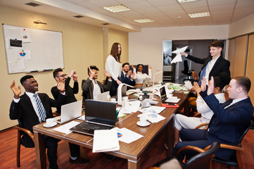 Multiracial business team meeting around boardroom table, two team leaders throw paper up.