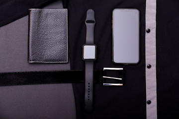 Top view of accessory on black shirt background. Set of black belt, smartwatch and smartphone. Composition of business man set. Copy space for text.