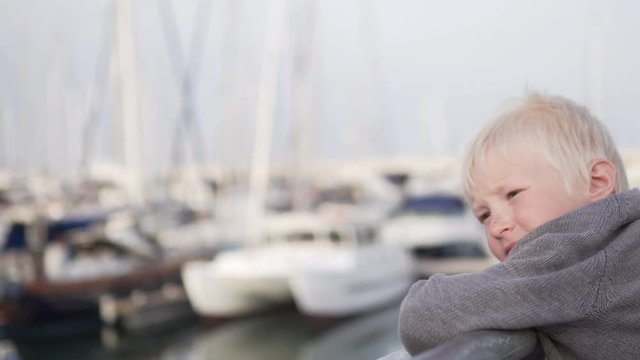 A sad blond boy stands on the pier near the yachts and looks at the sea. The child is waiting for his boat near the shore.