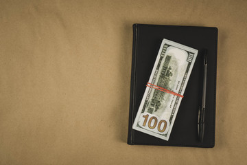 A black notebook , a pen and dollar cash banknotes on wooden background - concept of financial management or planning, make money from freelance writing