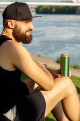Happy man with beard sits on the bank of the river in black cap, black T-shirt and black shorts and holds green can of beer