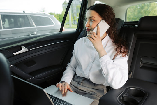Female entrepreneur travelling to office in a luxurious car sitting on backseat with laptop and looking outside the window while talking on phone. - Powered by Adobe