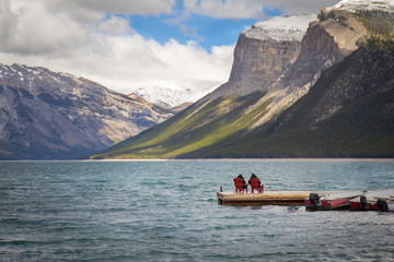 A couple sitting in red chairs on a pier in the middle of lake Minnewanka, Banff National Park, Canada, Rocky Mountains in the background