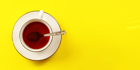 Tabletop view - white porcelain cup with freshly brewed hot black tea, spoon, on yellow board lit by morning sun, banner space for text right