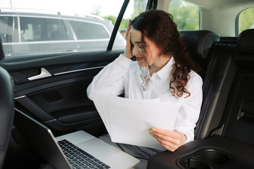 Upset confused business woman reading bad-news message, working with documents and laptop in a car.