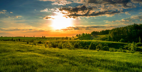 Fototapeta premium Magnificent scenery, sunset over fields, ravines and forests, turquoise orange sky and bright green grass and leaves of trees