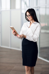 Attractive young woman wearing glasses and reading her touchscreen tablet while standing inside...