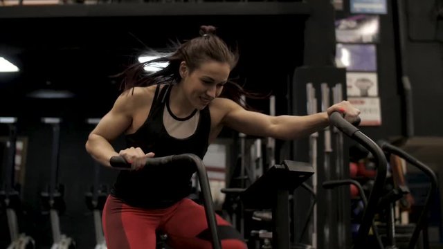 Sports woman is training on the stationary air bike and smiling in gym