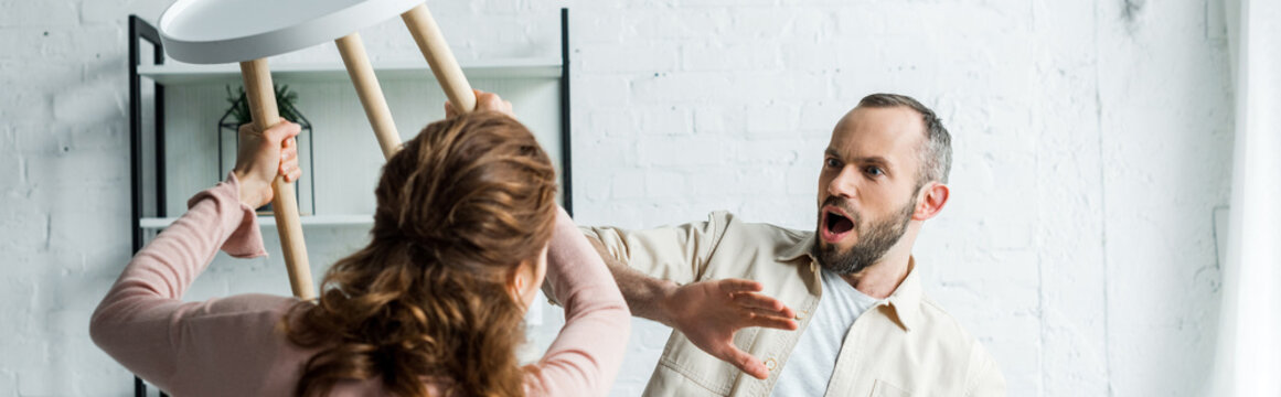 Panoramic Shot Of Surprised Man Looking At Angry Holding Chair While Threatening At Home