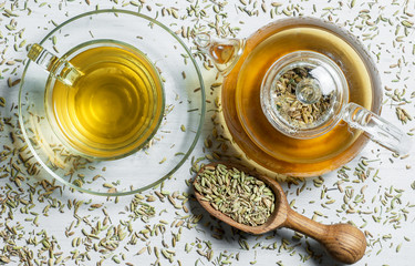 Herbal infusion fennel tea in glass cup and glass tea pot with dried fennel seeds in wooden shovel on white rustic table. Herbal tea alternative medicine background concept.