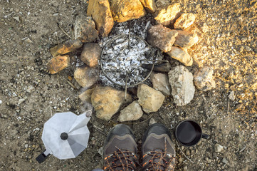 Preparing coffee using a bonfire during a camp in the countryside. First person view: bonfire with stones and ash, hiking boots, coffee maker and cup.