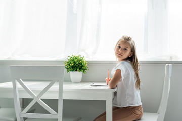 cute kid sitting and holding pencil near notebook at home