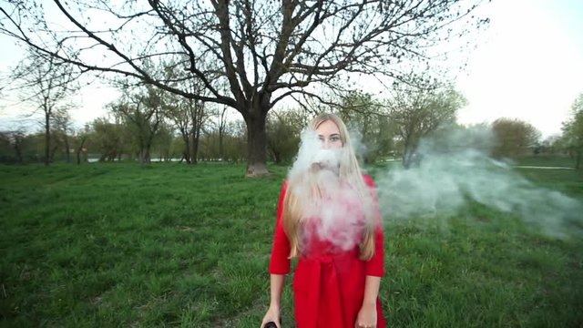 Vape woman. Young beautiful blonde girl in red dress smokes an electronic cigarette opposite a big tree in the evening outdoors in a park in spring. Bad habit that is harmful to health.