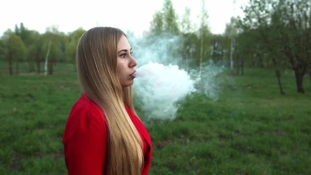 Vape woman. Young beautiful blonde girl in red dress and sunglasses smokes an electronic cigarette at sunset outdoors in a park in summer. Bad habit that is harmful to health. Vaping activity.