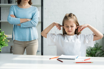 cropped view of woman standing with crossed arms near kid covering ears at home