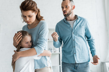 Fototapeta premium upset mother hugging daughter while angry man showing fist at home