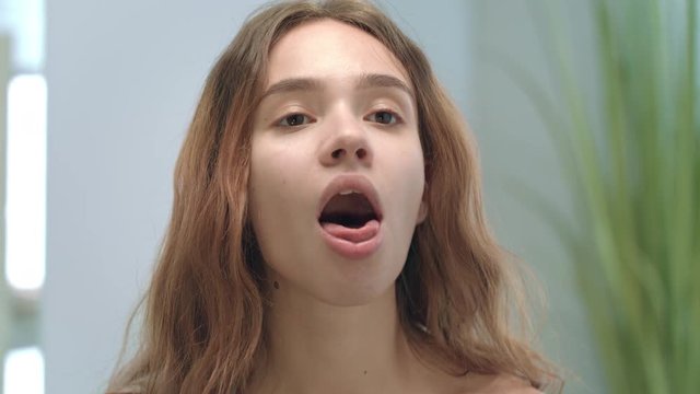 Young woman with opened mouth looking tongue and throat in mirror in bathroom