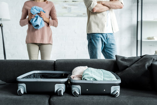 cropped view of man and woman standing near suitcase with clothes on sofa
