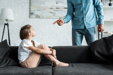 cropped view of man pointing with finger at upset kid sitting on sofa at home